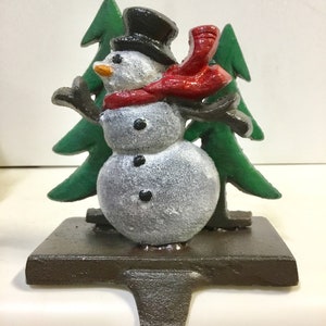 May include: A cast iron Christmas stocking holder featuring a snowman with a black top hat, red scarf, and a carrot nose. The snowman is set against two green Christmas trees. The base is dark brown with a hook for hanging.