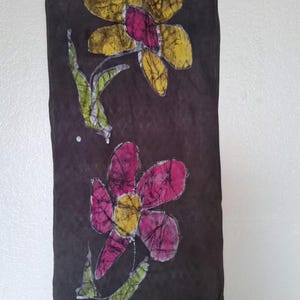 May include: A dark brown scarf with a floral design. The flowers are yellow and pink with green leaves. The scarf has a fringe at the bottom and is hanging on a gray hanger.