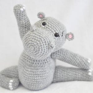 Puede incluir: Un juguete de peluche de hipopótamo gris tejido a crochet sentado con una pierna levantada en una pose de yoga. El hipopótamo tiene orejas rosas y ojos negros.