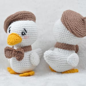 May include: Two white crocheted duck plushies wearing brown berets and brown bow ties. The ducks are facing different directions.