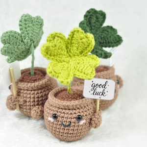 Puede incluir: Plantas de trébol de la suerte hechas a ganchillo en macetas marrones con caras sonrientes. Cada planta tiene un trébol verde de cuatro hojas y un letrero que dice "good luck". Las macetas tienen brazos y piernas.