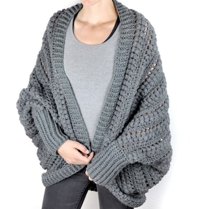 Könnte beinhalten: Ein grauer Häkelcardigan mit lockerer Passform und langen Ärmeln. Der Cardigan hat ein strukturiertes Stichmuster und einen weiten, offenen Ausschnitt.
