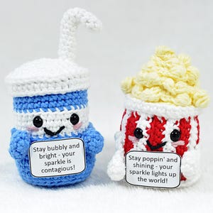 Könnte beinhalten: Zwei gehäkelte Plüschtiere, eines in Form eines Sodaglases mit Strohhalm und das andere in Form eines Popcorn-Eimers. Beide Spielzeuge haben lächelnde Gesichter und halten Schilder mit Botschaften über ihren Glanz. Das Sodaglas ist blau und weiß, und der Popcorneimer ist rot und weiß.