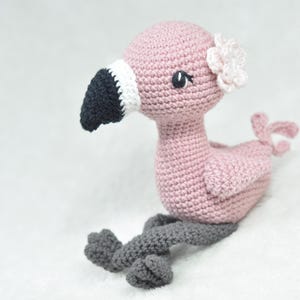 May include: A handmade crochet flamingo in shades of pink, grey, black, and white. The flamingo has a black beak with white trim, grey legs, and a small flower on its head. The bird is sitting on a white surface.