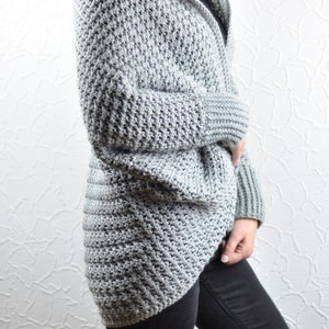 Könnte beinhalten: Ein grauer Kapuzenpullover mit einem strukturierten Stichmuster. Der Pullover hat lange Ärmel und eine lockere Passform.