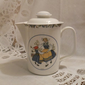 May include: White ceramic teapot with a blue and green floral design around the top. The teapot has a handle and a lid. The front of the teapot features a colorful illustration of two girls in traditional dress dancing.