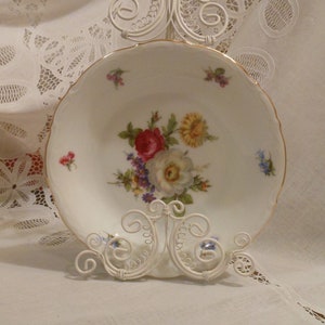 May include: A white porcelain bowl with a gold rim and floral design. The bowl is on a white metal stand.