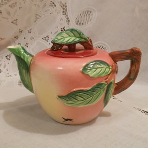 May include: A pink ceramic teapot shaped like an apple with green leaves and a brown handle.
