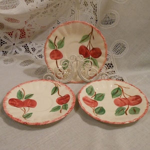 May include: Three white ceramic saucers with a red and green cherry design. The saucers have a scalloped edge.