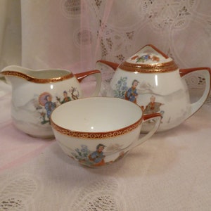 May include: A vintage porcelain tea set featuring a teapot, creamer, and teacup. The white pieces have a red and gold trim and are decorated with a colorful Asian-inspired scene. The teapot has a matching lid.