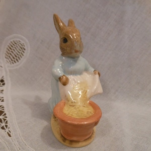 May include: A ceramic figurine of a brown rabbit wearing a blue dress, pouring yellow seeds from a white bag into a brown pot.
