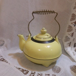 May include: A yellow ceramic teapot with a black stripe around the top and a spring-loaded handle. The teapot is sitting on a white lace tablecloth.
