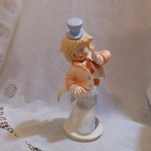 May include: A porcelain figurine of a clown with blonde hair wearing a blue hat and orange and white clothing. The clown is holding a hand to its ear and has a painted-on smile.