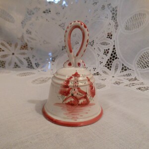 May include: A white ceramic bell with a red handle and a painted scene of a red building with trees. The bell has a red rim.