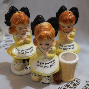 May include: Set of three vintage ceramic salt and pepper shakers featuring praying girls with yellow dresses and black bows. The shakers have different sayings: "We give you thanks for all your gifts", "We give you thanks for all your gifts", and "Give us this day our daily bread".