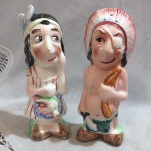 May include: A pair of vintage ceramic figurines depicting Native American figures. One figure wears a headdress and has an eye patch. The other figure wears a headband and a striped dress. Both have painted facial features.