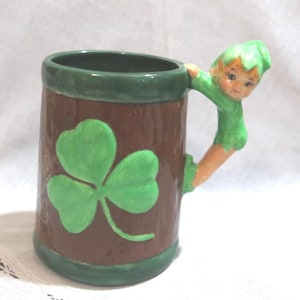 May include: A ceramic mug with a brown exterior and a green rim and base. A large green shamrock is painted on the front. The handle is a green leprechaun figure. A charming St. Patrick's Day themed mug.