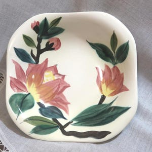 May include: A square, off-white ceramic dish with rounded corners. The dish is decorated with hand-painted pink and yellow flowers, green leaves, and dark brown stems. The floral design is centered on the dish.