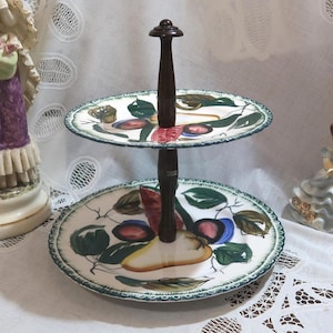 May include: Two-tiered serving tray with a dark wooden handle. The ceramic plates feature a hand-painted design of fruits and green leaves. The plates have a scalloped edge with a green trim. Decorative figurines are in the background.