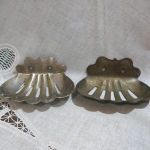 May include: Two vintage, silver-toned metal soap dishes. Each dish is shaped like a scallop shell with a scalloped edge and vertical slits. The dishes are displayed on a white, lace-trimmed surface.