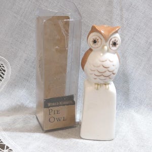 May include: A ceramic owl figurine with brown and white coloring, perched on a white rectangular base. The owl has detailed eyes and feather markings. The figurine is next to a clear plastic box with a brown cardboard label that reads "World Market Pie Owl."
