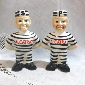 May include: A pair of ceramic salt and pepper shakers shaped like smiling figures in black and white striped prison uniforms. The word "ALCATRAZ" is printed in red on the chest of each figure. One figure has an "S" on its hat, the other has a "P".