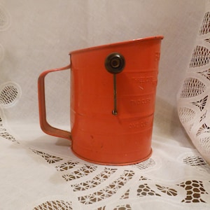 May include: A vintage red metal measuring cup with a handle and a swing-arm indicator. The cup has markings for one, two, and three cups. The handle is attached to the side of the cup. The cup is sitting on a white lace tablecloth.