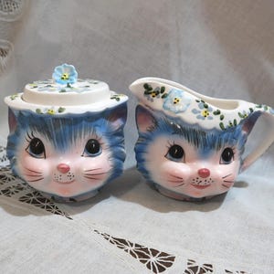 May include: A ceramic sugar bowl with a lid and a matching creamer pitcher, both shaped like cat heads. The pieces feature blue and white fur patterns, pink noses, black whiskers, and hand-painted blue flowers along the top rims.