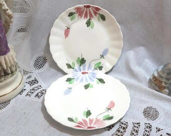 May include: Two white ceramic plates with scalloped edges. Each plate is decorated with a floral design in pink, blue, and green. The plates are on a white lace tablecloth.