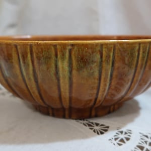May include: A brown ceramic bowl with vertical ribbed detailing. The bowl has a glossy finish with variations of brown and green hues. The bowl is likely vintage and suitable for serving or display.