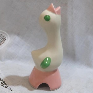 May include: A ceramic figurine of a white bird with a long neck, a pink beak, and green eyes and wings. The bird is perched on a solid pink base with a curved cutout at the bottom. The surface of the figurine is smooth and glossy in texture.