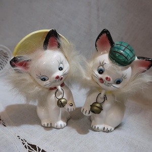 May include: A pair of white ceramic cat figurines with fluffy white fur. One cat wears a yellow hat and the other wears a green and white plaid hat. Both cats have bells around their necks.
