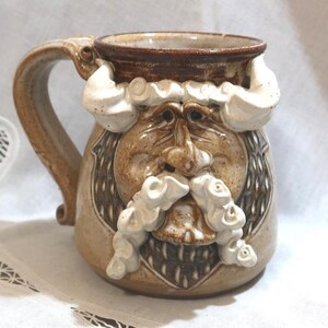 May include: A handcrafted ceramic mug with a sculpted face design. The mug features a tan base color with brown and white accents, including a beard and mustache. The handle is a matching tan color. The mug is designed to resemble a bearded man.