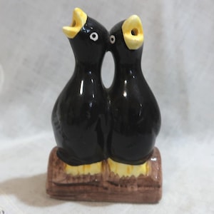 May include: A ceramic figurine of two black birds with open yellow beaks, perched on a brown base. The birds are facing each other, with white eyes and yellow feet. The figurine is likely a decorative item.