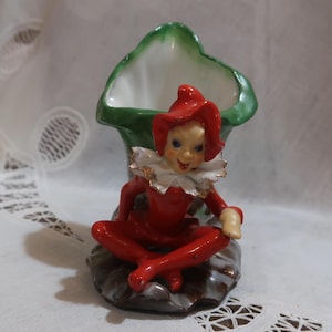May include: A red and white ceramic figurine of a jester sitting on a green and brown leaf-shaped planter. The jester is wearing a red and white hat and a red and white costume.