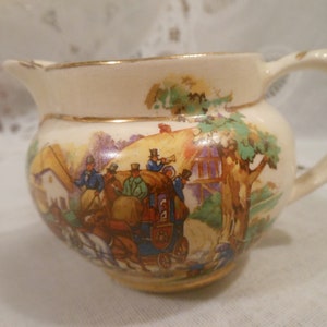 May include: A cream-colored ceramic creamer with a gold rim and a colorful scene of a horse-drawn carriage with people in it. The scene is painted on the side of the creamer.