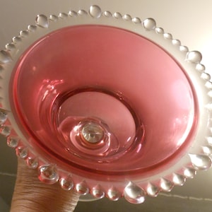 May include: A pink glass pedestal dish with a clear beaded edge. The dish has a scalloped rim and a small, round base.