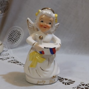 Puede incluir: Una figurita de porcelana blanca de un ángel con cabello amarillo y un halo. El ángel sostiene un tambor decorado con rayas rojas, blancas y azules. La base de la figurita está inscrita con "July 4th".