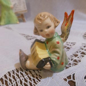 May include: A ceramic figurine of a seated angel playing an accordion. The angel wears a green dress with red polka dots and has tan hair. The accordion is yellow and black, and the angel has colorful wings.