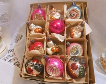 May include: A box of vintage Christmas ornaments with a variety of designs, including a pink, red, and blue ornament with a white figure, a silver ornament with a white figure, and a green and gold ornament with a red figure.
