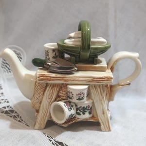 May include: A decorative teapot with a garden theme. The teapot is cream-colored with a wooden-style base and a green basket on top. It features miniature gardening tools and floral-decorated cups. The handle and spout are curved.