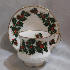 May include: A white teacup and saucer with a gold rim and a Christmas holly and pine cone design. The cup and saucer are decorated with green holly leaves, red berries, and brown pine cones.