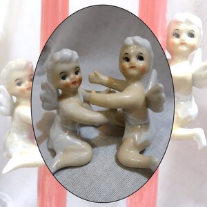 May include: Two porcelain cherub figurines with white wings and light brown bodies. The cherubs are holding hands and are sitting on a white surface.
