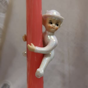 May include: A white porcelain figurine of an elf climbing a pink pillar. The elf is wearing a white hat and has a whimsical expression.