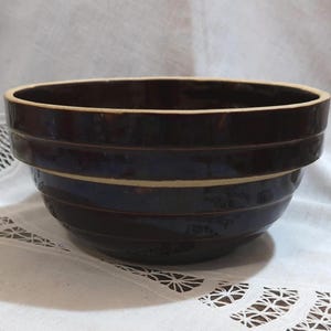 May include: A dark brown ceramic bowl with a glossy finish. The bowl has a wide, round shape with horizontal bands and a light beige rim. It sits on a white, patterned surface.