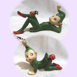 May include: A ceramic elf figurine in two poses, featuring a green suit, red shoes, and a gold-tipped hat. The elf has a pale face with large eyes and is accompanied by a small red and black ladybug perched on its body in both positions.