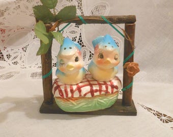 May include: Two ceramic blue and yellow bird figurines sit atop a picnic basket with a red and white checkered cloth. The birds are positioned within a small wooden frame structure decorated with green leaves and a thin green ribbon.