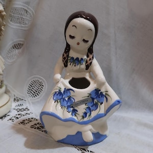 May include: A white ceramic figurine of a girl with brown hair and a blue and white dress. The dress is decorated with blue flowers and the word "Panchita" is written on the dress. The figurine has a small hole in the top of her head, suggesting it may be a planter.