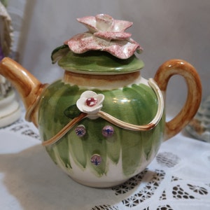 May include: A green and white ceramic teapot with a floral design. The teapot has a brown handle and a lid with a pink flower on top.