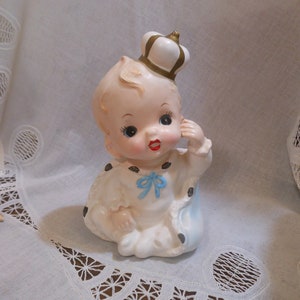 May include: A ceramic figurine of a baby wearing a white crown with gold trim and a blue and white outfit. The baby has a painted face with red lips and blue eyes.